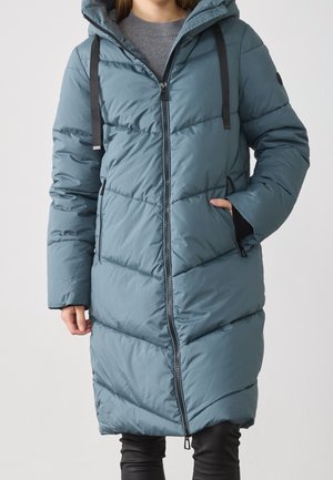 Femme portant un long manteau matelassé bleu sarcelle avec une fermeture éclair devant, une capuche avec des cordons noirs, un pull gris et un pantalon noir, debout avec une main dans la poche.