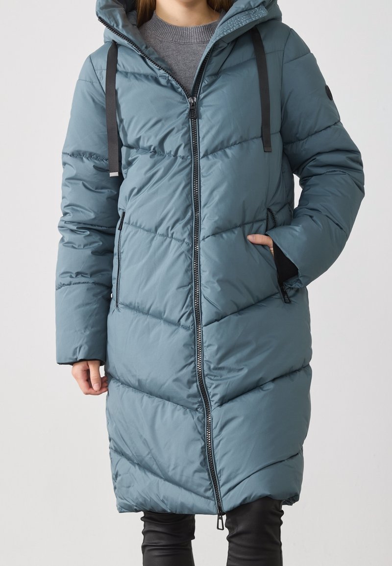 Femme portant un long manteau matelassé bleu sarcelle avec une fermeture éclair devant, une capuche avec des cordons noirs, un pull gris et un pantalon noir, debout avec une main dans la poche.