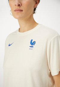 Kerman värinen puuvillapaita, jossa on sininen Nike-logo ja brodeerattu kukko-embleemi "FFF" alla. Lyhyet hihat, pyöreä kaula-aukko.