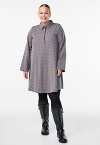 Nainen hymyilee harmaassa pitkähihaisessa mekossa, jossa on mustia pilkkuja, mustissa sukkahousuissa ja mustissa polvipituisissa saappaissa, seisten valkoista taustaa vasten.