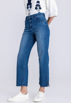 Vrouw in blauw recht gesneden jeans met zijsplitten, gecombineerd met witte sneakers en een wit shirt met een grafische zonnebrilmotief.