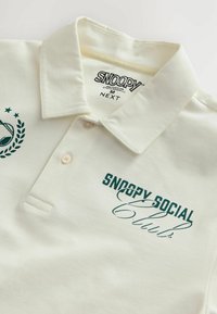 Crème poloshirt van katoen met een opgerolde kraag. Bevat groene tekst en logo "SNOOPY SOCIAL Club" op de borst. Twee knopen zichtbaar.