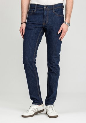 Mężczyzna ubrany w ciemnoniebieskie, dopasowane dżinsy, białe trampki z szarymi paskami, białą koszulę, zegarek na rękę i bransoletkę, stojący na jasnym tle.