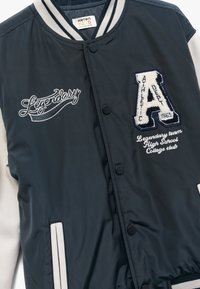 Granatowa kurtka varsity z kremowymi rękawami, zatrzaskami oraz haftowanym napisem "Legendarny" i logo "A". Gładki, teksturowany materiał oraz pasiakowy kołnierz.