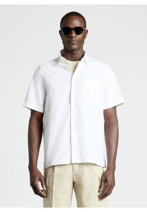 Uomo con occhiali da sole neri, camicia bianca a maniche corte con bottoni, pantaloni beige, in piedi davanti a uno sfondo grigio chiaro, rivolto in avanti.