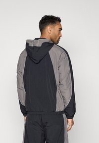 Chaqueta puffer negra y gris con capucha, que presenta detalles de ribete claros. Tejido texturizado con puños y dobladillo elásticos, proporcionando un ajuste relajado.