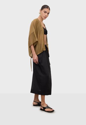 Woman wearing a loose brown kimono-style jacket over a black crop top, long black skirt, and black open-toe sandals, standing against a gray background.
