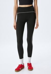 Mustat leggingsit korkealla vyötäröllä, joissa on valkoiset sivu raidat ja sileä pinta. Yhdistettyinä punaisiin lenkkareihin ja valkoisiin lyhyisiin sukk main.