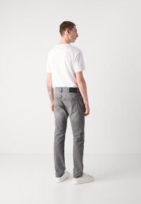 Jeans de mezclilla grises para hombre de corte recto, con bolsillos traseros y una etiqueta negra en la cintura, combinados con una camiseta blanca sencilla y zapatillas blancas.