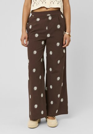Pantalon marron à jambes larges avec motifs de soleil blancs, porté avec des espadrilles beiges et un haut court en maille crème sur une personne debout.