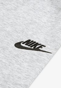 Svetlo siva bombažna tkanina z črno vezeno Nike logotipom z značilnim zamahom. Tekstura izgleda mehka in gladka.
