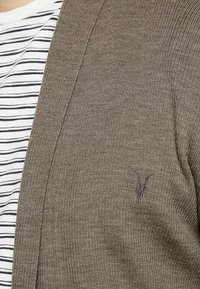 Cardigan marrone lavorato a maglia con scollo a V, caratterizzato da un piccolo logo scuro sul petto sinistro. Indossato sopra una camicia a righe bianche e blu navy.