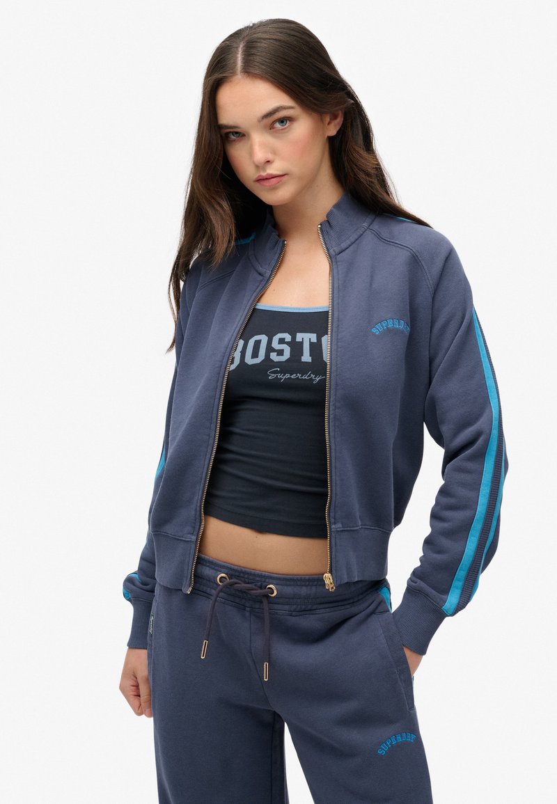 Sudadera con cremallera azul marino, con rayas azul claro, diseño ajustado, puños acanalados y un logo. Combinada con un conjunto atlético azul marino a juego.