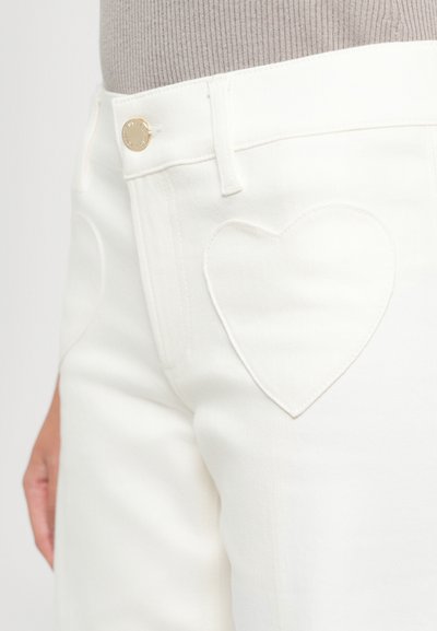 Pantalons blancs avec une poche en forme de cœur cousue sur la cuisse avant, fermeture à bouton doré, portés avec un haut en rib de couleur beige clair.