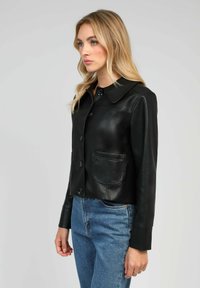 Veste en cuir noir avec un col, des boutons à l'avant, deux poches poitrine et des manches longues. Portée sur un jean en denim bleu. Texture lisse.