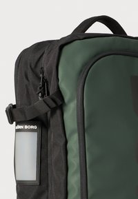 Ryggsekk med et mørkegrønt og svart design. Har en teksturert overflate, en sidelomme, og et rektangulært logoskilt på forsiden.