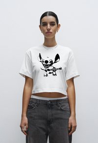 Bílé zkrácené tričko s černým motivem postavy hrající na kytaru. Látka působí měkce, s krátkými rukávy a volným střihem.