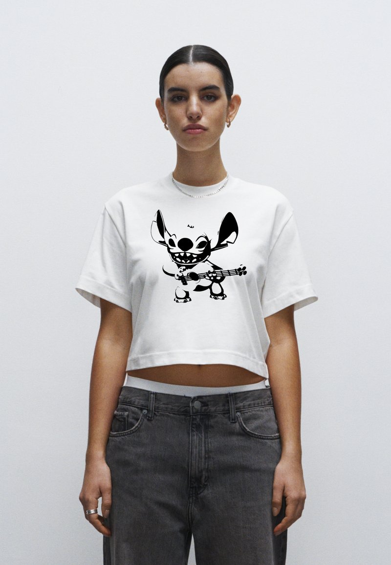 Bílé zkrácené tričko s černým motivem postavy hrající na kytaru. Látka působí měkce, s krátkými rukávy a volným střihem.