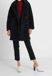 Abrigo negro de piel sintética con doble botonadura, grandes solapas y botones negros, combinado con pantalones de rayas finas y mules blancos de punta abierta.