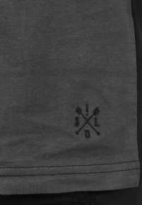 Mørkegrå bomulds T-shirt-stof med en glat tekstur, der har et sort broderet logo af krydsede pile ved den nederste kant.