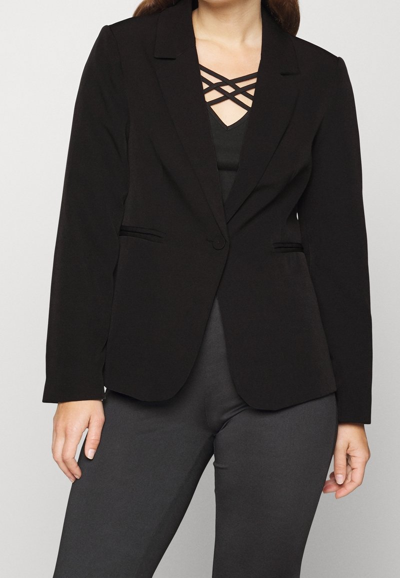 Femme portant un blazer noir cintré sur un top noir à bretelles croisées, associé à un pantalon gris foncé sur un fond clair uni.