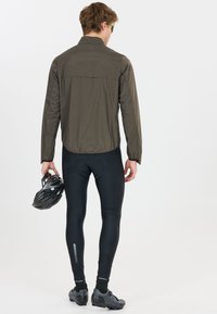 Homme en veste olive et collants de cyclisme noirs tenant un casque noir, vu de dos sur fond blanc.