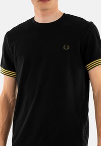 Zwart katoenen t-shirt met korte mouwen, voorzien van gele en groene gestreepte accenten op de mouwen en een klein geborduurd logo op de borst.