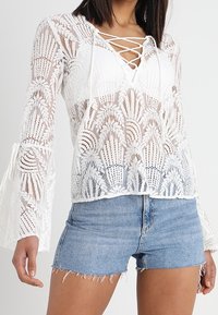Blusa bianca in pizzo con scollatura a V e laccio frontale, caratterizzata da maniche a campana e motivi floreali. Abbinata a pantaloni shorts in denim.