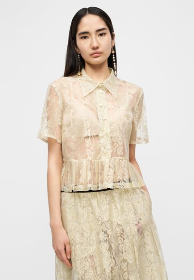 Δαντελένιο μπλουζάκι σε απαλό παστέλ κίτρινο με κοντά μανίκια, μυτερό γιακά, κλείσιμο με κουμπιά και πλισέ τελείωμα. Διαφανής υφή με λουλουδάτο μοτίβο.