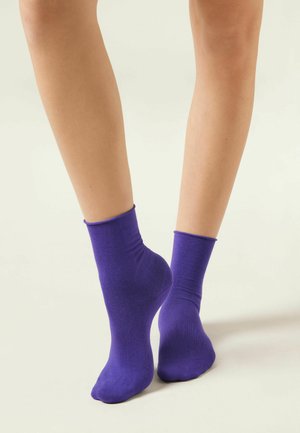 Par de piernas desnudas con calcetines tobilleros morados, con un pie ligeramente levantado sobre las puntas, sobre un fondo claro y liso.