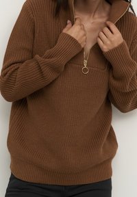 Personne portant un pull en maille épaisse marron avec col zippé, tenant les bords du col, habillée d'un pantalon noir sur un fond neutre.