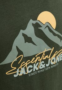 T-shirt en coton vert foncé avec un design de montagne gris et un soleil jaune, portant le texte "Essentiels JACK & JONES" en blanc.