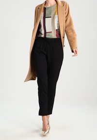 Cappotto cammello su un maglione geometrico multicolore, abbinato a pantaloni neri e tacchi metallici, con una vestibilità su misura e un design elegante.