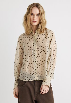 Femeie blondă cu păr ondulat, purtând o bluză bej cu nasturi și buline negre și pantaloni largi maro închis, stând pe fundal alb.