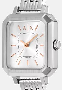 Reloj de pulsera cuadrado de plata con marcadores de hora en oro rosa, tres manecillas y una correa de eslabones metálicos en un primer plano.