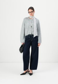 Grijze fluffy cardigan met knopen aan de voorkant, donkerblauwe wijde jeans, zwarte puntige hakken en een witte sjaal met stippen om de hals.