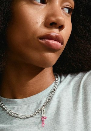 Zilveren ketting met een roze "liefde" hanger, gedragen over een lichtgrijs T-shirt. De ketting heeft een robuust schakeldesign.