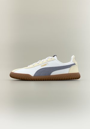 White sneakers featuring a grey suede stripe, yellow accents, and a textured brown rubber sole. Laced with light grey shoelaces.