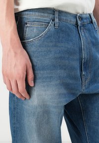 Nærbillede af en persons hånd, der hviler ved siden af blå, falmede denim jeans med synlige syninger og lomme detaljer.