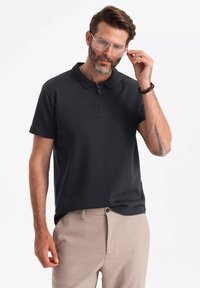 Tmavě šedé polo tričko s krátkým zipem u límce, vyrobené z hladké tkaniny. Skombinováno se světlými béžovými kalhotami, které mají volný střih.