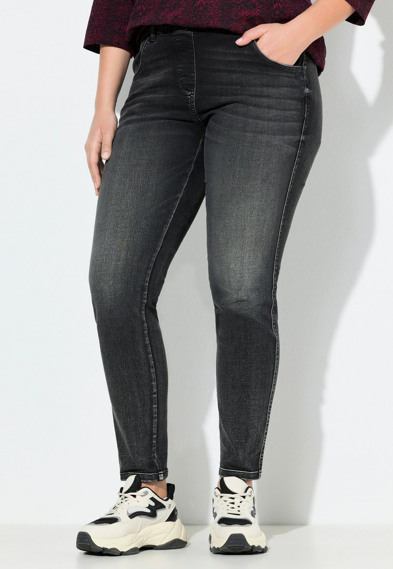 Personne portant un jean skinny noir délavé et des baskets blanches épaisses, avec une main dans la poche, sur un fond blanc uni.
