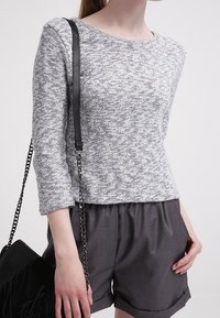 Person, der ein graues meliertes Langarmoberteil und dunkelgraue Shorts mit aufgeschlagenem Saum trägt, mit einer schwarzen Schultertasche mit Kettenriemen.