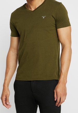 Mężczyzna noszący oliwkowozielony t-shirt z krótkim rękawem i dekoltem w serek, z małym białym logo GANT po lewej stronie klatki piersiowej, w połączeniu z czarnymi spodniami.