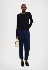 Svart knapp-up cardigan, marineblå bukser, blanke sorte loafers og en liten hvit håndveske. Jevn tekstur, tettsittende design og diskret logo-detalj.