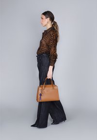 Tan lederen handtas met gestructureerd ontwerp, bovenhandvatten en een kleurrijke sleutelhangeraccent. Model in een shirt met luipaardprint en donkere broek.