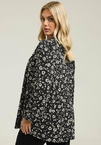 Blusa nera con motivo floreale bianco, maniche lunghe, vestibilità ampia, orlo arrotondato e tessuto texturizzato. Nessun hardware o accentuazione notevoli visibili.