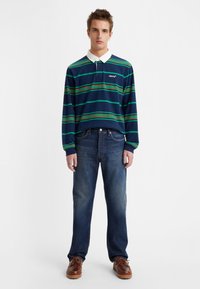 Polo majica s dolgimi rokavi v mornarsko modri črti z zelenimi in rdečimi črtami, bela ovratnica, v kombinaciji z modrimi denim hlačami in rjavimi vezalnimi čevlji.