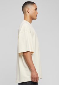 Beige løstsittende bomulls t-skjorte med korte ermer, rund hals og myk tekstur, vist fra siden mot en nøytral bakgrunn.