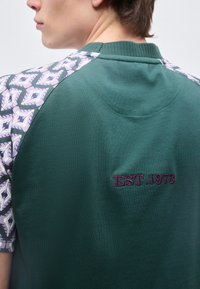 Νεαρό άτομο που φοράει πράσινο πουκάμισο με μοτίβο σε μωβ μανίκια και κεντημένο το "EST.1978" στην πλάτη κοντά στον ώμο.