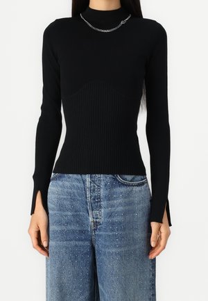 Femme portant un haut noir ajusté à manches longues côtelées avec des fentes pour pouce aux poignets et un jean taille haute bleu orné de petites décorations blanches.